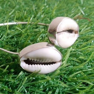 Op de afbeelding: Een zilverkleurige armband met twee grote, witte kaurischelpen. De schelpen zijn bevestigd op een dunne, gebogen band. De armband ligt op groen gras.