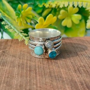 Könnte beinhalten: Ein silberner Spinner-Ring mit drei Edelsteinen: Türkis, Mondstein und blauer Topas. Der Ring hat ein strukturiertes Band und ist auf einer Holzoberfläche mit verschwommenem floralen Hintergrund platziert.