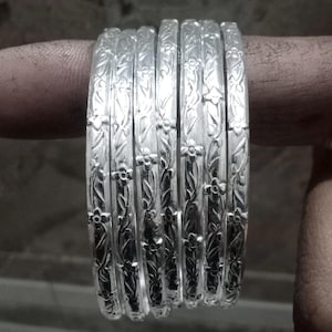 May include: A set of seven silver bangles. Each bangle features a floral and leaf pattern. The bangles are stacked together, showcasing the intricate design. The silver reflects light, highlighting the details of the engraved pattern.