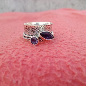 May include: A silver spinner ring with floral detailing and two purple gemstones. One gemstone is oval, the other is marquise-cut. The ring is set on a pink surface.