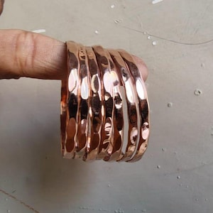 Op de afbeelding: Een stapel van zes koperkleurige armbanden met een gehamerde textuur. De armbanden zijn rond en hebben een glanzend, reflecterend oppervlak. De armbanden zijn op elkaar gestapeld en tonen hun ontwerp en kleur.