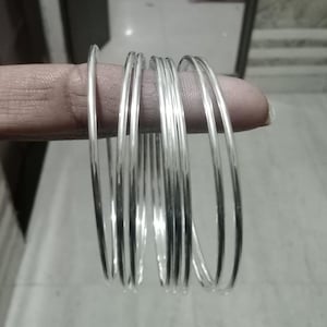 May include: A collection of thin, silver-colored bangles. The bangles are round and appear to be made of metal. The image shows several bangles stacked together, reflecting light.