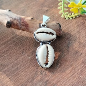 May include: A silver pendant featuring two white cowrie shells. The shells are set in a silver frame and connected, with a silver loop at the top for a chain. The pendant is displayed on a wooden surface.