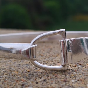 Könnte beinhalten: Ein polierter Silberarmreif mit einem zentralen Verschluss in Steigbügelform. Die Metalloberfläche ist glatt und reflektierend und zeigt ein durchgehendes Banddesign, das an der Vorderseite mit einem markanten, vom Reitsport inspirierten Element verbunden ist.