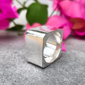 Könnte beinhalten: Ein dicker, polierter Silberring mit einem markanten quadratischen Band. Die Metalloberfläche ist glatt und reflektierend und zeigt eine solide, klobige Konstruktion mit abgerundeten Ecken und einer hohlen Mitte in einem klaren Finish.