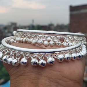 May include: A pair of shiny silver-toned metal bangles featuring a smooth, rounded band design. Each bangle is adorned with a series of small, spherical jingle bells suspended from the lower edge, creating a decorative, dangling effect.