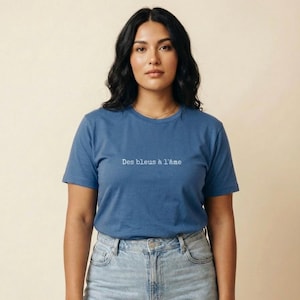 May include: A blue t-shirt with the phrase "Des bleus à l'âme" printed in white. The shirt has short sleeves and a crew neck. The model is wearing blue jeans.