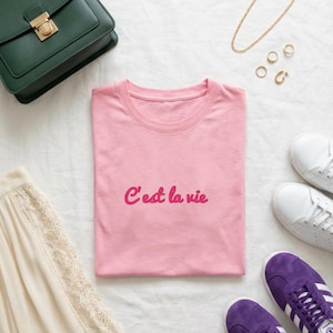 Op de afbeelding: Een roze t-shirt met de tekst "C'est la vie" in roze. Het shirt is omgeven door accessoires, waaronder een groene handtas, gouden sieraden, witte sneakers en paarse sneakers.