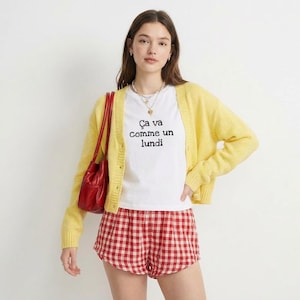 Peut inclure: Une femme porte un cardigan jaune, un t-shirt blanc avec le texte "Ça va comme un lundi" et un short à carreaux rouges et blancs. Un sac à main rouge est suspendu à son épaule. Elle porte également un collier.