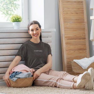 Puede incluir: Una mujer con una camiseta gris oscuro con la palabra "COMPOSED" en blanco, sentada en una alfombra. Sostiene una cesta de mimbre llena de ropa doblada. La escena incluye una lavadora y una ventana.
