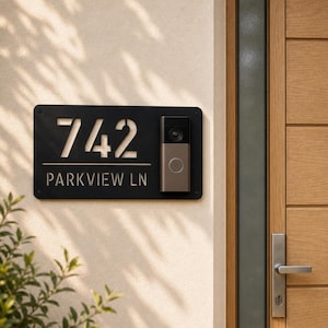 May include: A rectangular black metal house sign featuring the cut-out numbers 742 and the text PARKVIEW LN, with a mounted video doorbell integrated on the right side, secured to a light-colored wall next to a wooden door.