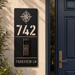 May include: A rectangular black metal address plaque featuring a compass rose graphic at the top, the house number 742, and the text PARKVIEW LN at the bottom. A dark-toned video doorbell is mounted in the center of the vertical metal plate.