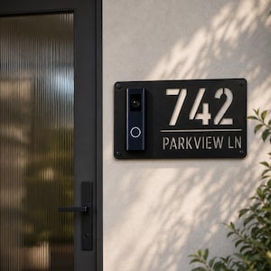 May include: A rectangular black metal house number plaque featuring the number 742 and the text PARKVIEW LN, with a vertical black video doorbell mounted on the left side. The plaque is secured to a light-colored wall with four visible screws.