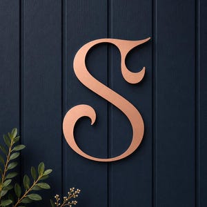 May include: A copper-colored metal letter S with a decorative, swirling serif font. The letter features a smooth, matte finish and is mounted against a dark navy blue vertical paneled wall, with a small green leafy plant visible at the bottom left.