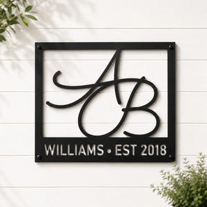 May include: A square black metal wall sign featuring a large, stylized script monogram of the letters A and B centered within a frame. Below the monogram, the text WILLIAMS • EST 2018 is cut out in a clean, sans-serif font. Four screws secure the sign.