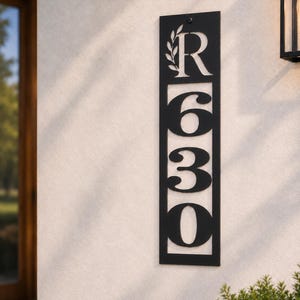 May include: A vertical rectangular black metal house number sign featuring the letter R at the top accompanied by a leaf sprig design, followed by the numbers 6, 3, and 0 stacked vertically. The sign is mounted to a light-colored wall surface.
