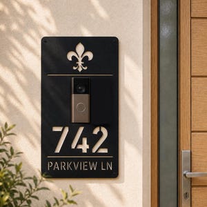 May include: A rectangular black metal house sign featuring a cutout fleur-de-lis symbol at the top, a central mounted video doorbell, the house number 742, and the text PARKVIEW LN laser-cut into the surface, mounted on a light-colored wall.