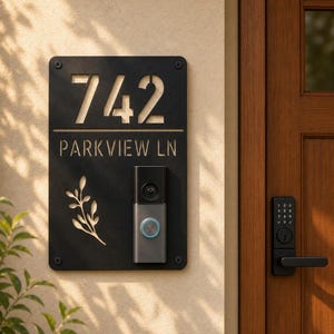 May include: A rectangular black metal house number sign featuring the cutout text 742 and PARKVIEW LN, along with a decorative leaf motif. A grey video doorbell with a circular blue light is mounted on the lower right side of the sign.