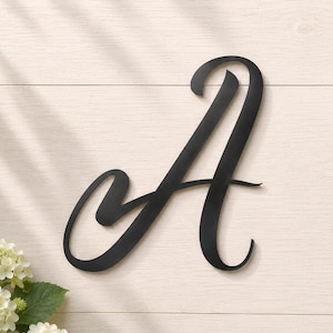 May include: A matte black metal wall decoration in the shape of the uppercase letter A. The letter features a flowing, cursive script font with smooth, continuous lines and a flat finish. The design is simple, clean, and lacks any additional embellishments.