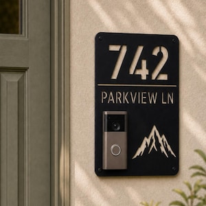 May include: A rectangular black metal house sign featuring the laser-cut numbers 742 and the text PARKVIEW LN. A silver video doorbell is mounted on the lower left side, and a mountain range graphic is cut out on the lower right side.