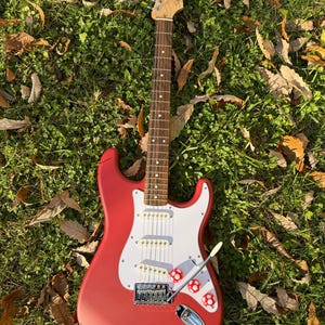 May include: A red electric guitar with a white pickguard and mushroom-shaped knobs. The guitar has a wooden neck and headstock, and is resting on a bed of green grass and fallen leaves.