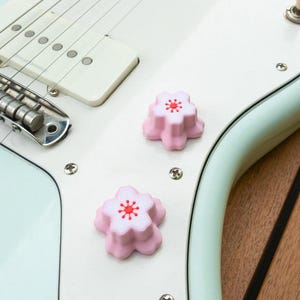 May include: Close-up of a light blue electric guitar with white pickguard. Two pink flower-shaped knobs with red centers are on the pickguard. The guitar's strings and pickups are visible. The background is a wooden surface.