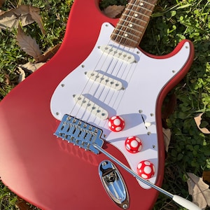 May include: A red electric guitar with a white pickguard and three white pickups. The guitar features three red and white mushroom-shaped knobs. The guitar is resting on a bed of green grass and fallen leaves.