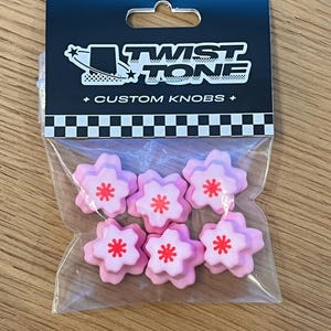 May include: A package of six pink flower-shaped custom knobs with red centers. The package has a black header with the text "TWIST TONE + CUSTOM KNOBS +" and a checkered pattern.