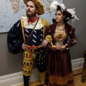 May include: A couple dressed in Renaissance-era clothing. The man is wearing a yellow and blue doublet, yellow breeches, and a brown hat with a feather. The woman is wearing a burgundy dress with gold trim and a white feathered hat.