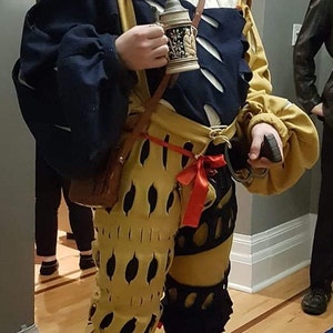 May include: A person wearing a brown and gold costume with a large, wide-brimmed hat. They are holding a ceramic mug with a floral design. The costume features a red sash and black and gold patterned pants.