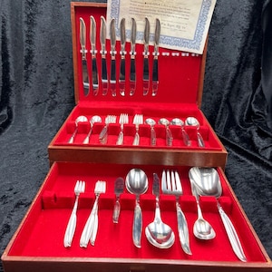 Puede incluir: Un cofre de cubiertos de madera con forro de terciopelo rojo, abierto para revelar un juego completo de cubiertos plateados. El juego incluye cuchillos, tenedores, cucharas y utensilios para servir, todos ordenados.