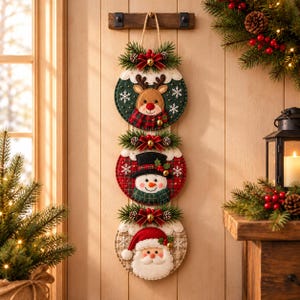 May include: A vertical hanging Christmas decoration featuring three circular felt ornaments stacked together. The top ornament shows a reindeer, the middle a snowman, and the bottom a Santa Claus, each accented with pine sprigs, red bows, and bells.