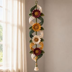 May include: A vertical hanging wall decoration featuring a central green cord with four crocheted flowers in maroon, mustard yellow, and cream. The flowers are interspersed with spiraling, open-weave ribbons in cream, green, and yellow, finished with a tassel.