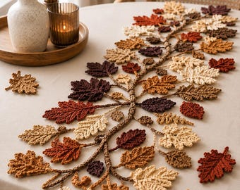 May include: A crochet table runner featuring a central vine with numerous attached leaves in shades of cream, tan, orange, brown, and dark purple. The leaves are arranged in a sprawling pattern across a light-colored tablecloth, with several loose leaves scattered nearby.