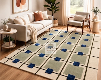 May include: A rectangular area rug featuring a grid pattern with large light sage green squares, smaller dark blue square accents, and a cream-colored background. The rug displays a logo with the text The Artisan Rug Crafts in the center.