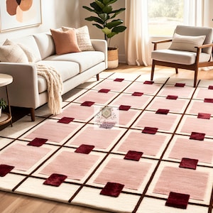 Könnte beinhalten: Ein großer Teppich mit geometrischem Design. Der Teppich hat einen cremefarbenen Hintergrund mit einem Gittermuster aus dunkelbraunen Linien. Innerhalb des Gitters befinden sich hellrosa Rechtecke und dunkelrote Quadrate. Der Teppich liegt in einem Wohnzimmer.
