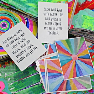 May include: A collection of colorful art pieces and instructions. White cards with text: "ADD GLOBS OF PAINT ON YOUR PAGE THEN COVER IT WITH PLASTIC WRAP AND SMOOSH IT DOWN. REMOVE AND LET DRY." and "SPRAY YOUR PAGE WITH WATER. DIP YOUR BRUSH IN WATER COLORS AND LET IT BLEED TOGETHER."