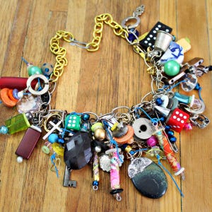 Puede incluir: Un collar de charms colorido con una variedad de abalorios, incluyendo dados, botones, llaves y una pequeña bobina de hilo. El collar está hecho de cadena de plata y tiene un acento de cadena dorada.
