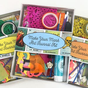 May include: A Make Your Mark Art Journal Kit in a rectangular box featuring a label with a heart, a skull, and the text Make Your Mark Art Journal Kit. The kit contains a roll of orange tape, a roll of pink lace, a glue stick, and various paper cutouts.