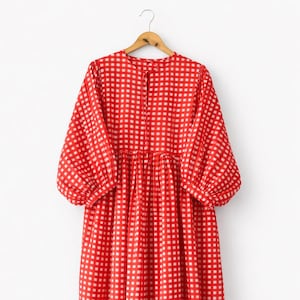 May include: A red and white gingham dress with long, puffed sleeves and a gathered waist. The dress is suspended on a wooden hanger. The fabric has a checkered pattern. The background is white.