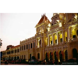Puede incluir: Un edificio grande y ornamentado con una fachada amarilla y un tejado de tejas rojas. El edificio tiene muchas ventanas y columnas, iluminadas por luces. La imagen se toma al anochecer, con el cielo amarillo pálido.