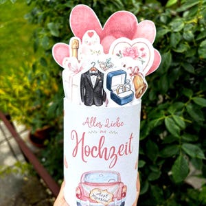 May include: A cylindrical wedding greeting card featuring the German text Alles Liebe zur Hochzeit. The design includes watercolor illustrations of a tuxedo, a ring box with two rings, a bell, a champagne bottle, and a pink car with a Just Married sign.