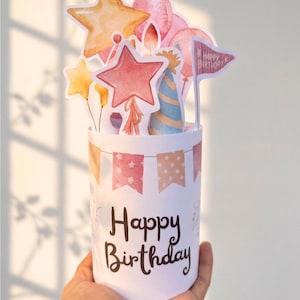 May include: A cylindrical white paper card featuring the text Happy Birthday in dark brown script. The top displays a pop-up arrangement of watercolor-style illustrations including yellow and orange stars, pink balloons, a blue party hat, and a flag.