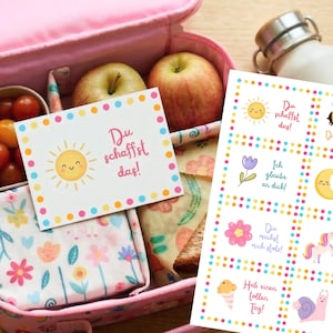 May include: A pink lunchbox containing cherry tomatoes, apples, and a sandwich. Inside is a card with a smiling sun and the text "Du schaffst das!". A sheet of stickers with illustrations and phrases is also visible.