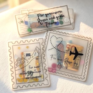 May include: Three translucent, rectangular envelopes designed like postage stamps and tickets, containing folded Euro banknotes and colorful confetti. The envelopes feature black printed text in German, including phrases like F&uuml;r gemeinsame Reisen und gro&szlig;e Tr&auml;ume, F&uuml;r euer erstes Abenteuer als Ehepaar, and F&uuml;r eure Reise ins Gl&uuml;ck.