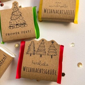 May include: Four small rectangular chocolate bars wrapped in brown kraft paper sleeves. Each sleeve features black ink illustrations of Christmas trees or gift boxes and German text including Frohes Fest, eine wundervolle WEIHNACHTSZEIT, and herzliche WEIHNACHTSGR&Uuml;BE. The sides show red, green, and yellow foil.