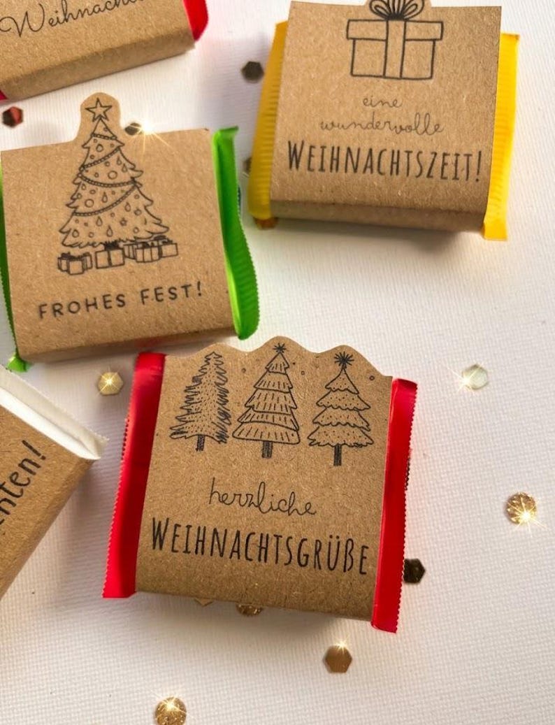 May include: Four small rectangular chocolate bars wrapped in brown kraft paper sleeves. Each sleeve features black ink illustrations of Christmas trees or gift boxes and German text including Frohes Fest, eine wundervolle WEIHNACHTSZEIT, and herzliche WEIHNACHTSGR&Uuml;BE. The sides show red, green, and yellow foil.