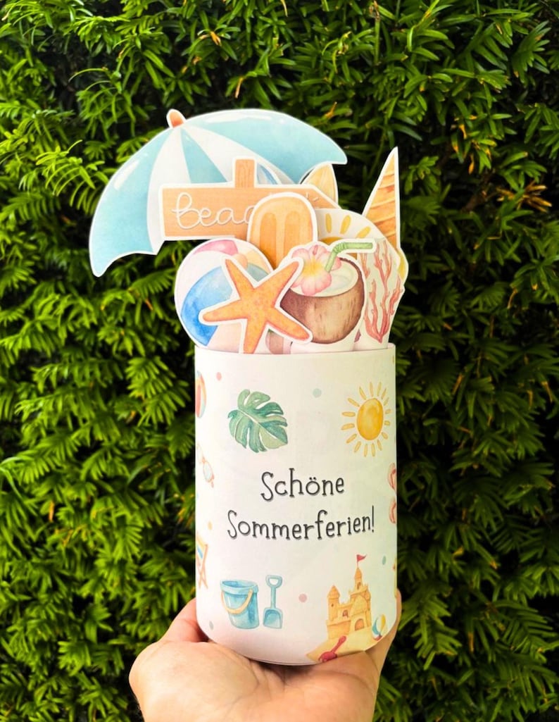 May include: A cylindrical container featuring the text "Sch&ouml;ne Sommerferien!" and summer-themed illustrations including a sandcastle, sun, and bucket. The top is decorated with cutouts of a beach umbrella, starfish, coconut drink, and ice cream.