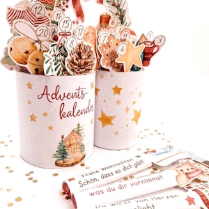 May include: Two white cylindrical containers decorated with gold stars and a house illustration, labeled Advents kalender. They hold various numbered Christmas-themed cutouts. Three white rectangular bars feature German text including Frohe Weihnachten.