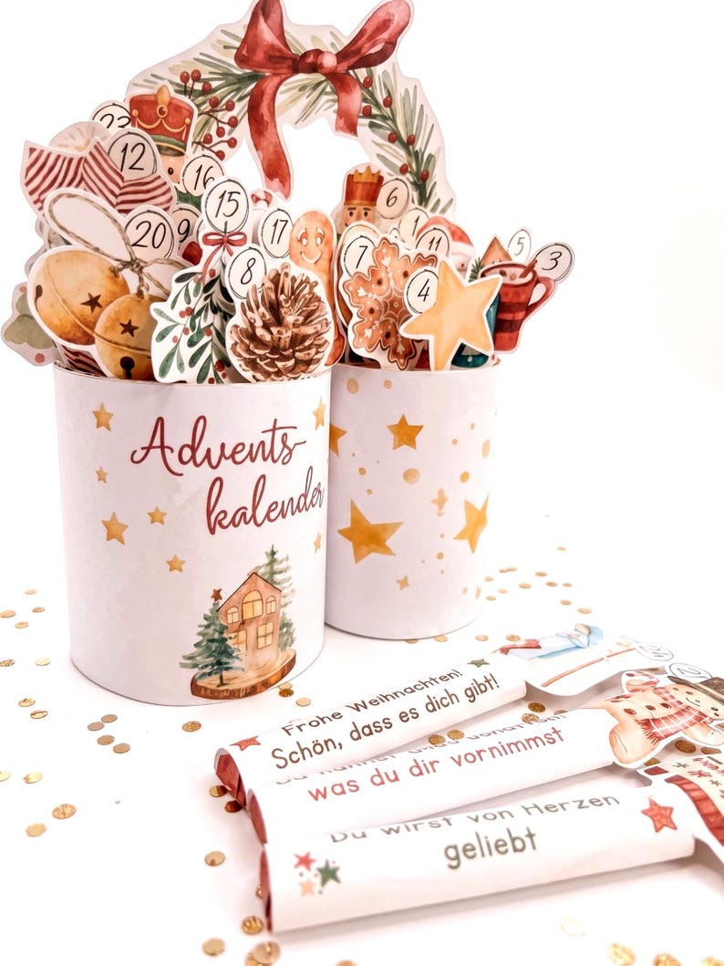 May include: Two white cylindrical containers decorated with gold stars and a house illustration, labeled Advents kalender. They hold various numbered Christmas-themed cutouts. Three white rectangular bars feature German text including Frohe Weihnachten.